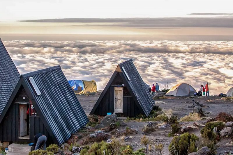 Wooden A-frame huts on a bare ridge above a sea of cloud.