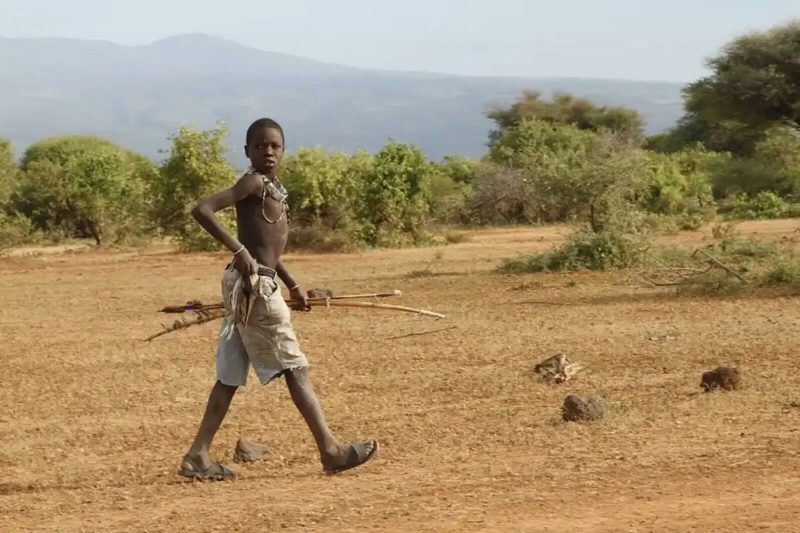 A boy carrying a bow and arrows walks across dry bush country at the foot of a hill.