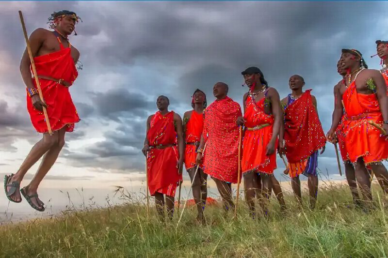 A Maasai man leaping into the air beside a line of others in red shukas.