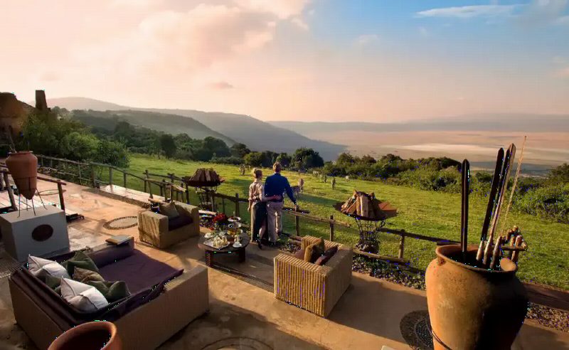 Guests on a lodge terrace with sofas and lanterns, looking out across a green valley at dusk.
