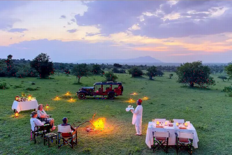Tables laid for dinner around a campfire in the bush at dusk, a safari vehicle parked behind.