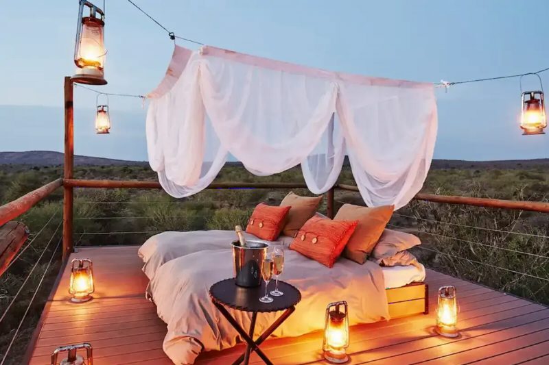 An outdoor bed under a white mosquito net on a wooden deck, hung with glowing lanterns at dusk.