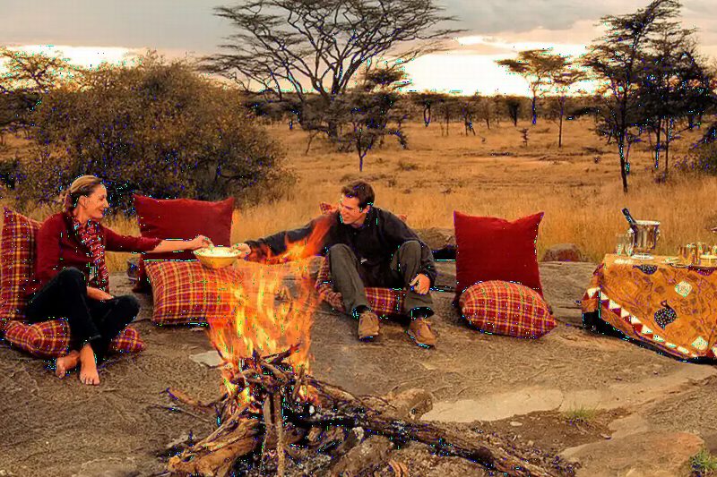 A couple sitting on red cushions beside a campfire in the bush.