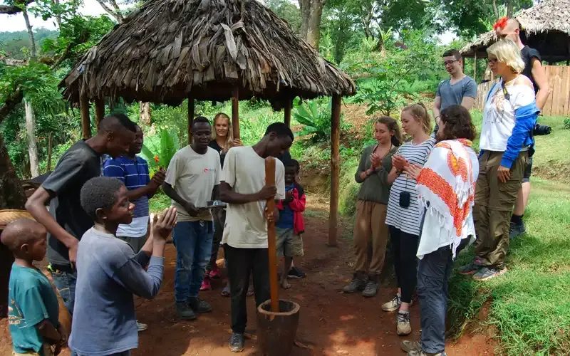 Visitors talking with villagers beside a thatched shelter in green, leafy country.